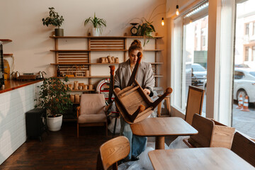 An adult White woman is setting up furniture inside a modern cafe. She arranges a wooden chair on a...