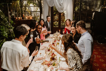 A multiracial group of young and adult men and women toast wine at a decorated indoor dinner table during an evening celebration. Several are wearing formal suits and dresses.
