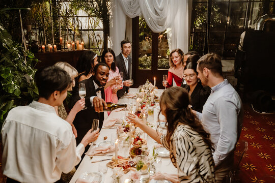A multiracial group of young and adult men and women toast wine at a decorated indoor dinner table during an evening celebration. Several are wearing formal suits and dresses.