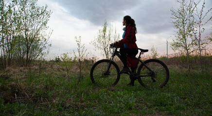Obraz premium Young woman riding a bicycle on the spring field with green grass on sunset sky landscape background. Happy activity sport trip vacations