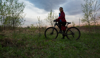 Young woman riding a bicycle on the spring field with green grass on sunset sky landscape background. Happy activity sport trip vacations
