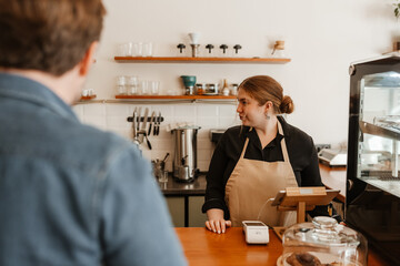 A white adult woman wearing an apron works at a cafe counter, assisting a male customer. The scene shows a moment inside a cozy, modern cafe environment with coffee equipment and food displayed.