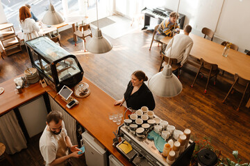 A barista interacts with a woman at the counter of a cozy cafe. The cafe features wooden furniture, a modern espresso machine, and several people sitting at tables enjoying drinks and conversation.