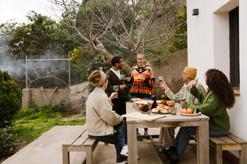 A multiracial group of adult men and women gather around a wooden table in an outdoor courtyard, wearing sweaters and sipping wine.