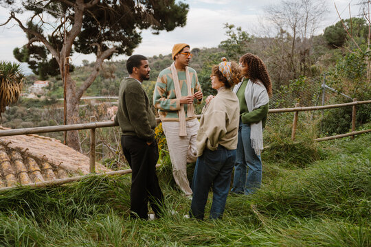 A multinational group of adult people is standing outdoors in casual sweaters and scarves, engaged in a lively conversation in a natural setting with trees and lush grass in the background.