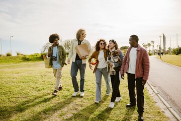 A multinational group of young people, a Black man, a White man, a Latina woman, an Asian woman, and a dog, walking together in a park, enjoying a picnic with a volleyball, camera, and picnic basket.
