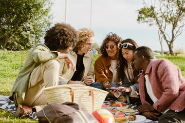 A group of young multiracial men and women sit on a blanket in a park, looking at a smartphone and enjoying a picnic with fruits and snacks on a sunny day.