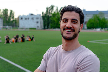Portrait of a smiling soccer coach standing with arms crossed on the field with his team resting in...