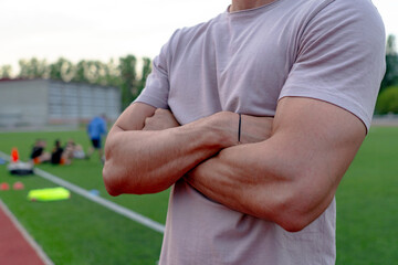 Coach standing with arms crossed beside a soccer field, observing his team as they train and...