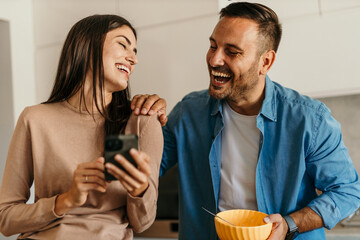 Happy couple laughing together while having breakfast and using smartphone in kitchen