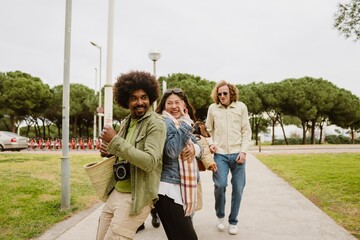 Obraz premium A group of young multiracial men and women walk outdoors on a park path, smiling and having fun, with a woman carrying a small dog on a cloudy day.