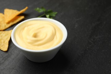Tasty dipping cheese sauce in bowl, nacho chips and parsley on black table, closeup. Space for text