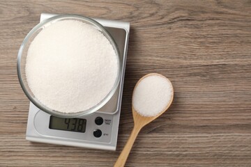 Digital kitchen scale with bowl of sugar and spoon on wooden table, flat lay. Space for text
