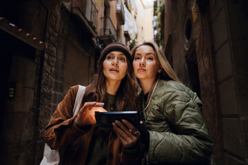 Two young White women stand closely together and use a smartphone for navigation. They explore an old, narrow street lined with historic stone buildings, wearing casual jackets and looking ahead.