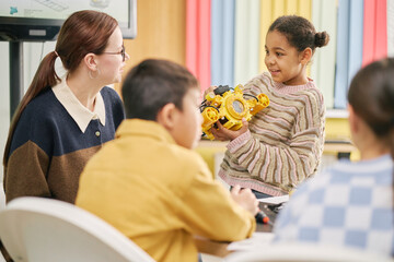 Young girl presenting robot to classmates in vibrant classroom environment featuring engaged teacher observing students interaction with curiosity