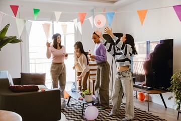 A group of diverse young women celebrate indoors at a casual party. They dance, hold drinks, and play with balloons under colorful flags in a bright apartment living room.