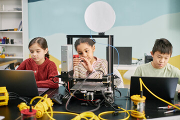 Children engaging in STEM activities in classroom with 3D printer and laptops, emphasizing hands-on learning and technology. Portrays concentration and collaboration among students