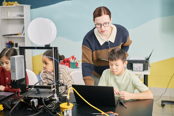 Teacher helping students with projects in technology classroom. Children engaged in learning with computers and 3D printer, emphasizing collaborative education environment