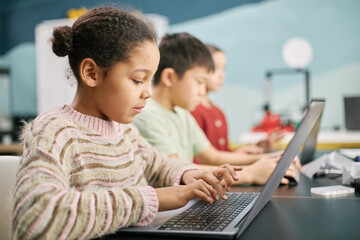 Multiracial children using laptops while sitting in row in classroom setting, focusing on learning and typing effectively © Seventyfour