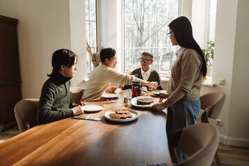 A family of Asian descent gathers around a wooden dining table, eating dessert and drinking tea in...