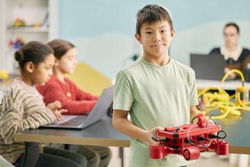 Portrait of boy holding robotic model, standing in classroom environment with focused students working on laptops in background