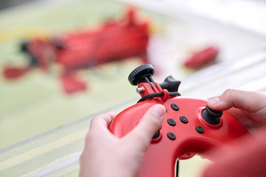 Close-up of hands holding a red gaming controller while engaging in gameplay. Background features a slightly blurred gaming setup creating a focused atmosphere