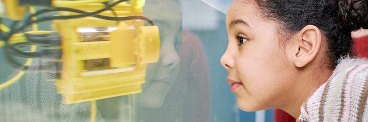 Website header shot of young girl of African descent closely observing a robotic device at a...