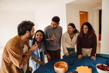 A group of young multiracial friends standing around the table eating snacks and laughing indoors at home.