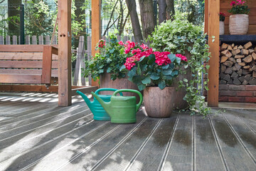 Green garden watering cans standing on the floor next to large planters with pink flowers in a summer gazebo in the garden