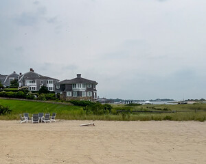 Elegant coastal homes overlooking a sandy beach with white lounge chairs and lush green grass, bordered by dunes and a calm waterfront under a soft, cloudy sky.