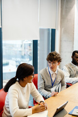 Male intern sitting at a desk holding clipboards while talking to a female employee sitting next to...