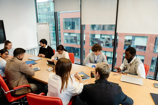 Group of eight employees and a male intern sitting at a table and talking, in an office