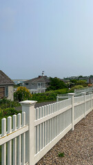 A peaceful summer scene featuring a white picket fence with a coastal view of boats, a distant beach, and charming beachside cottages under a clear sky.