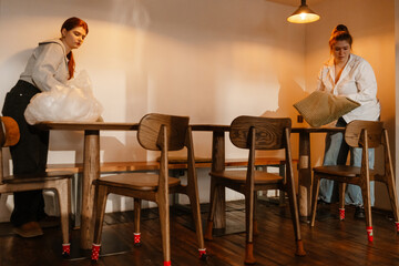 Female owner and female employee are standing at a table and holding a pillow and a bag, in a cafe