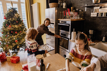 Mother smiles and opens the oven while her son holds a baking sheet with cookies next to her, in...