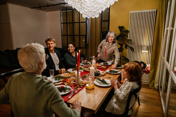 Family and guests sit at a table and smile while a grandmother stands next to them