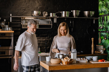 A man stands and looks at a table with a woman standing behind it, stirring a bowl with a whisk,...
