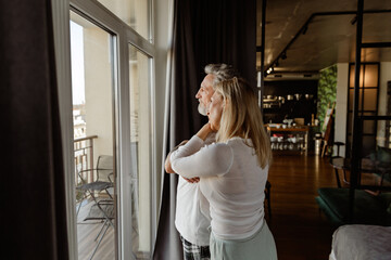 A woman hugs a man standing next to her, they look out the window