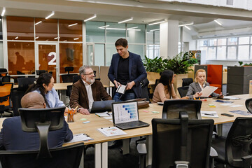A male employee stands and holds a notebook while a group of five employees sit at a table next to him, in a meeting