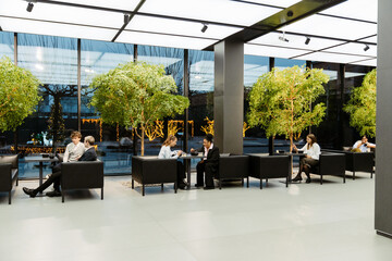 A group of five employees sit at tables and eat while a male intern sits next to one of them and...