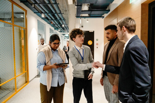 A male intern and a male employee stand and shake hands while two employees stand next to them, in a hallway