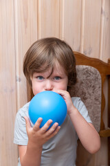 Blue-eyed boy blowing up a blue balloon while sitting on a wooden chair on a wooden background