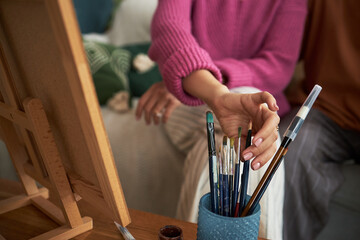 Close-up of woman choosing paintbrush to paint picture with her man in the room