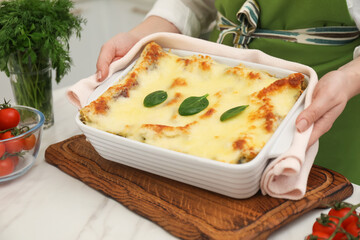 Woman putting baking dish with tasty spinach lasagna onto white marble table indoors, closeup