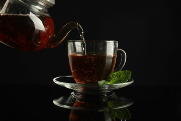 Pouring freshly brewed tea from teapot into cup at dark mirror table, closeup