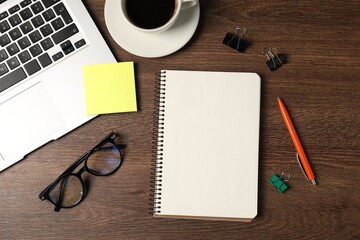 Notebook, pen, sticky note, binder clips, glasses, coffee and laptop on wooden table, flat lay