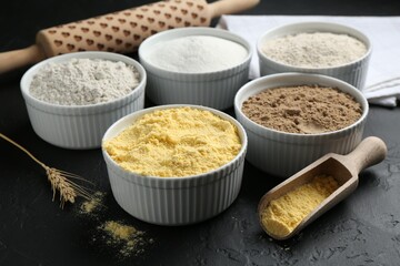 Different types of flour in bowls, scoop and rolling pin on black textured table, closeup