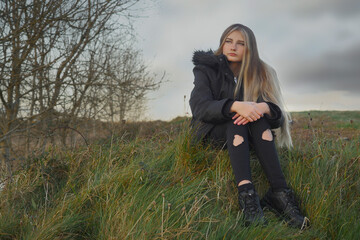 A girl is sitting on a hillside in the grass. She is wearing a black jacket and black pants, her...