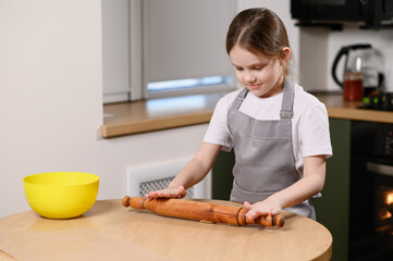 Portrait of cute little girl cooking in kitchen. Girl rolling out small piece of dough on kitchen...