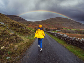 Teenager girl tourist in blue jeans and yellow hat and jacket walking on wet narrow road in a...